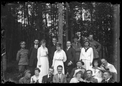 Grupa kobiet i mężczyzn na skaraju lasu. Część mężczyzn w mundurach wojskowych. Pierwszy po prawej na dole prawdopodobnie malarz Ludwik Pikor