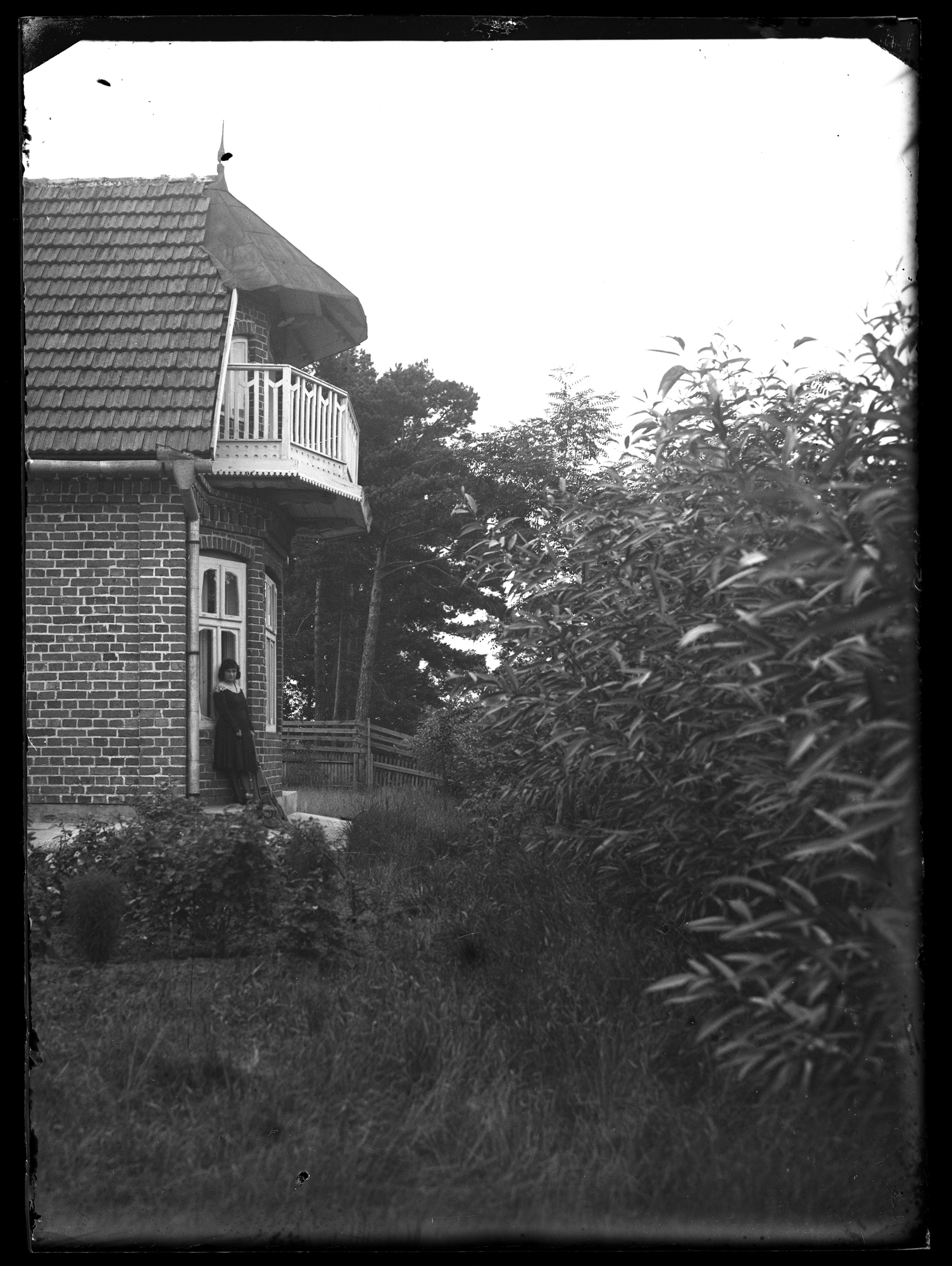 Fragment domu z ogrodem. Przed domem stoi kobieta
